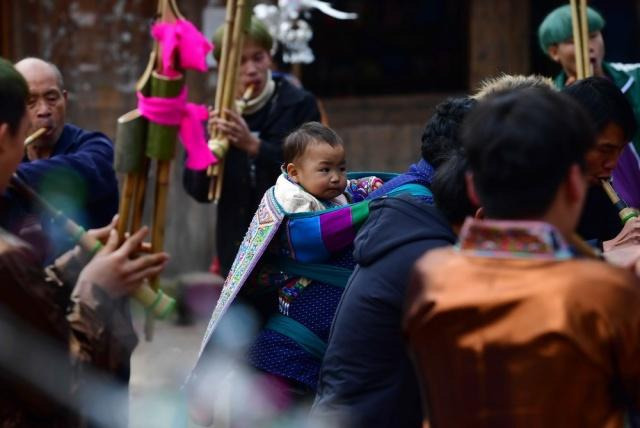 图为在乌英苗寨，村民背着小孩在吹芦笙。