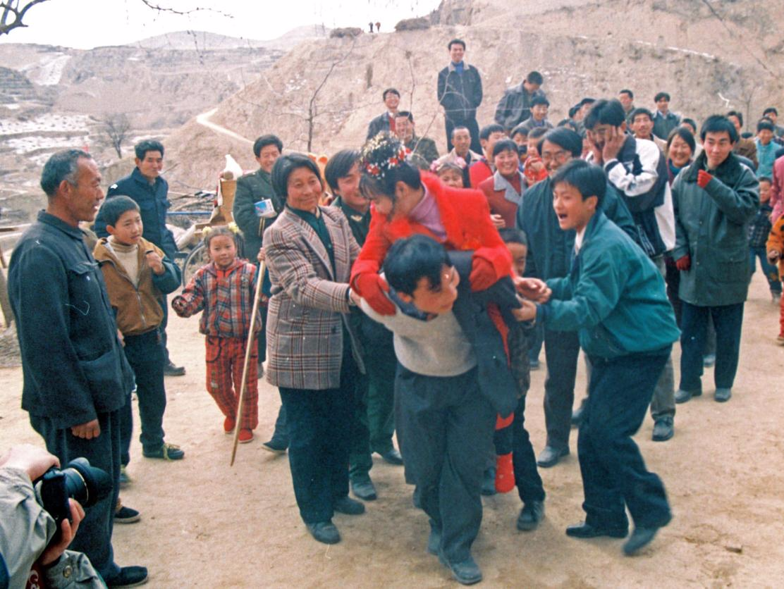 2000年春节，陕北米脂县刘峁村，鞭炮声中新郎在众人的拥簇下背着新娘走进了自家的大院。图片来源：姚忠智/视觉中国