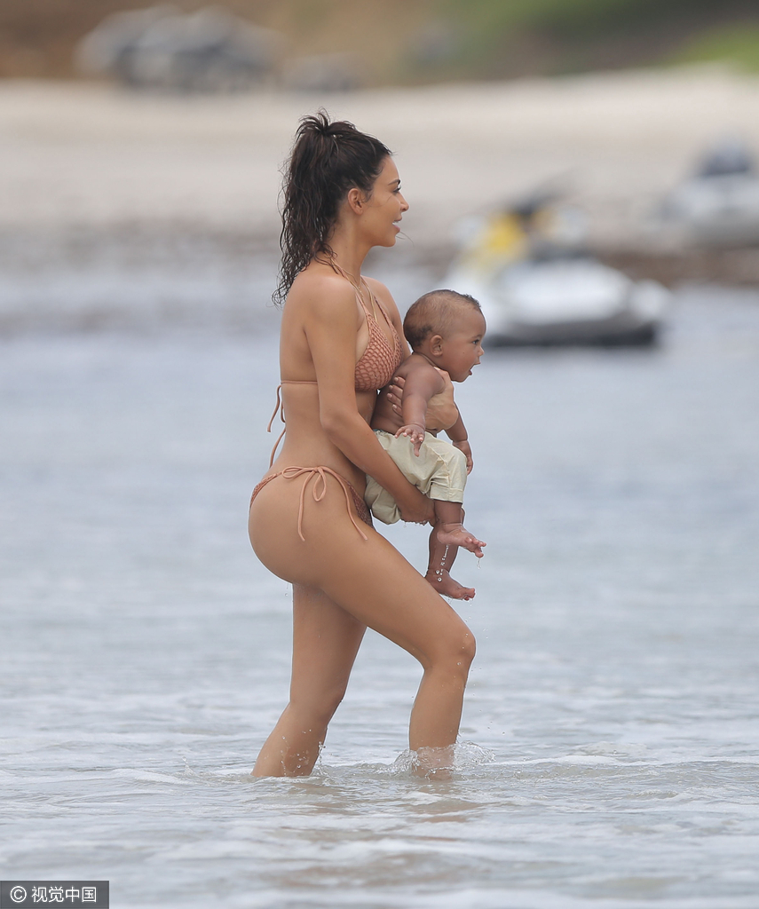 当地时间8月18日，金·卡戴珊（Kim Kardashian）带女儿小西北和儿子沙滩玩耍，卡戴珊身穿裸色系带比基尼，女儿与妈妈同款可爱十足，在沙滩上卡戴珊全程呵护幼子母爱爆棚，然而抢镜的还是翘臀。图为卡戴珊抱幼子。